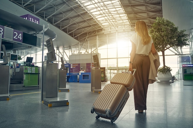 Attentive female person carrying her luggage and going to the necessary gate for taking her sit in plane