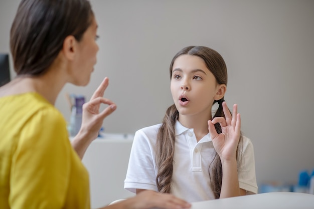 Attentive focused long-haired school-age girl repeating the sound and gesture of psychologist sitting opposite