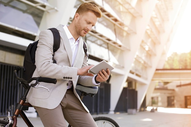 Uomo d'affari bello attento che utilizza una compressa e che si appoggia sulla bicicletta