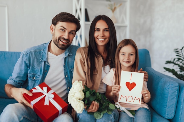 Attentive husband and loving child holding presents and flowers while celebrating mother day with a happy young woman