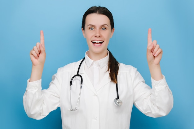 Attentive nurse, woman doctor in white coat and stethoscope, pointing fingers upper, posing over blue wall. Physician smiling amazed as shown awesome announcement. Healthcare workers, pandemic concept