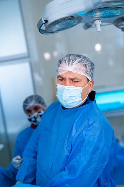 Attentive professional doctor checks operation process in modern operating room. Surgeon in blue scrubs, hat and facial mask in clinic. Portrait.