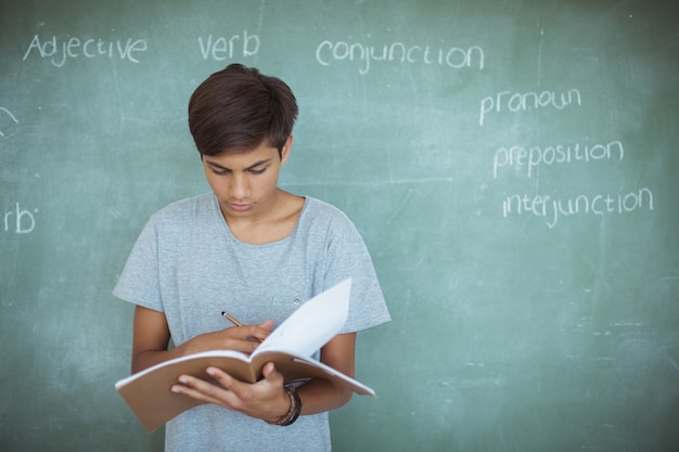 Attentive schoolboy reading book against chalkboard in classroom