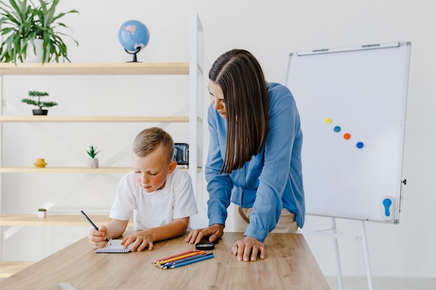 Attentive young woman tutor teacher helping little boy pupil with studying correct mistakes explain learning material Smiling mother assist small boy with home task