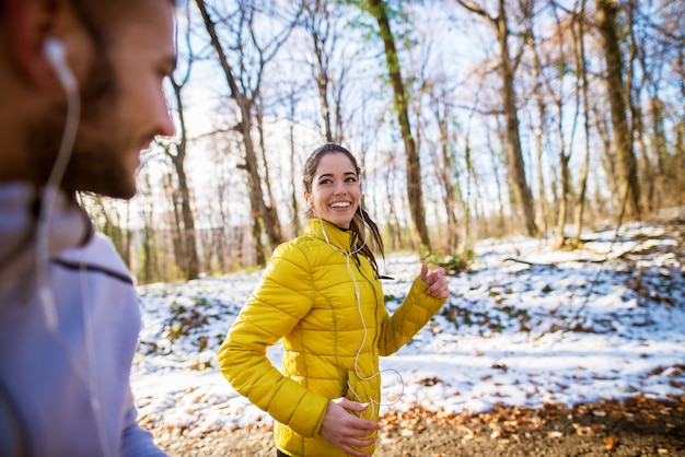 Attractiive улыбаясь подходит спортивная пара работает с наушниками через лес в зимнее утро.