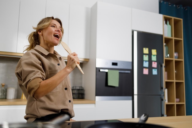 Attractive adult woman singing and dancing in the kitchen while holding a wooden spatula