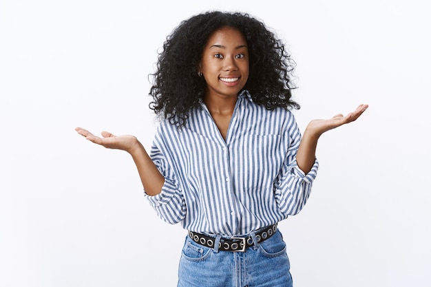 Attractive african-american curly-haired woman replying uncertain gesture shrugging raise hands sideways clueless, unable answer smiling sorry look, standing questioned uninvolved, white wall