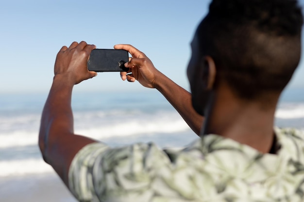 An attractive African American man enjoying free time on beach on a sunny day, standing on sand, using his smartphone, taking photos of himself.