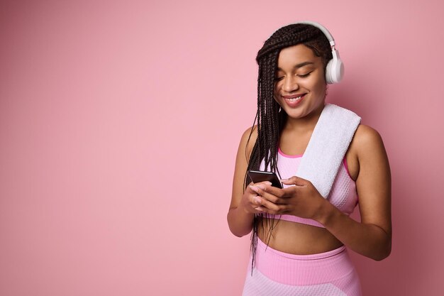 Photo attractive african american sportswoman in pink tracksuit and wireless headphones, browsing web pages on internet in smartphone, swiping, surfing, isolated over pink colored background