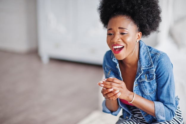 Attractive african american woman with afro hair wear on skirt and jeans jacket posed at white room Fashionable black model