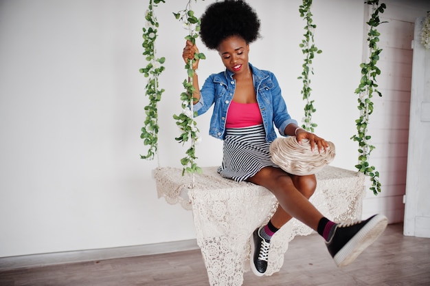 Attractive african american woman with afro hair wear on skirt and jeans jacket, posed at white room on swing. Fashionable black model.