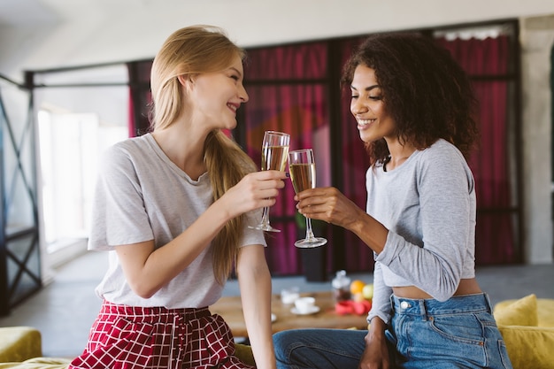 Attractive african american woman with dark curly hair and smiling woman with blond hair sitting on sofa holding glasses of champagne in hands while joyfully spending time together at home