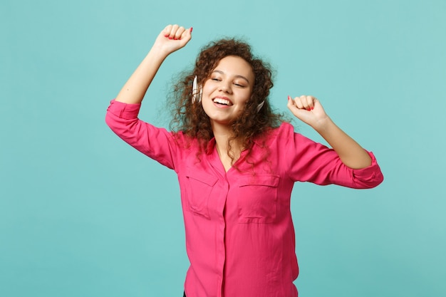 Attractive african girl in pink casual clothes listening music with headphones and dancing isolated on blue turquoise wall background. People sincere emotions, lifestyle concept. Mock up copy space.