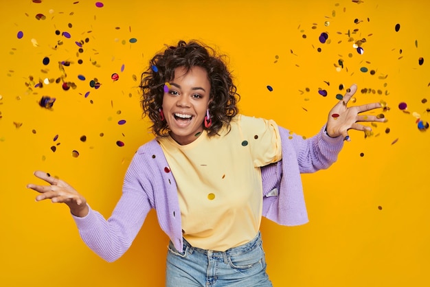 Photo attractive african woman throwing confetti and smiling while standing against yellow background