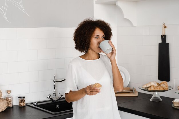 Привлекательная африканская женщина с кофе и пирожными на белой кухне Уютное утро афроамериканской девушки на кухне дома