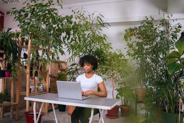 Attractive African young business woman working typing on laptop computer sitting at desk in home office room with modern biophilia design