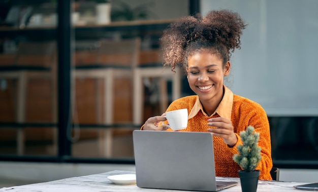 Attractive african young confident woman holding coffee cup and using laptop computer sitting at open coworkspace