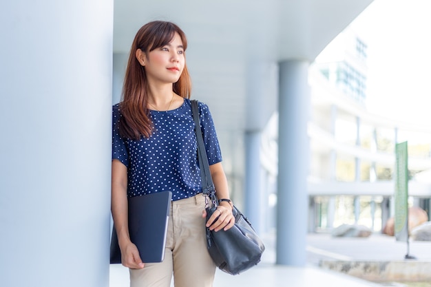 Attractive asian business woman standing leaning against building holding her computer and purse