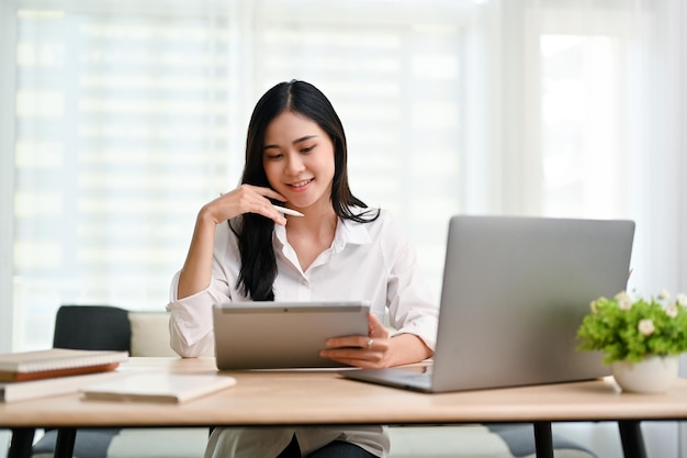 Attractive Asian businesswoman hand on chin looking at tablet screen at desk