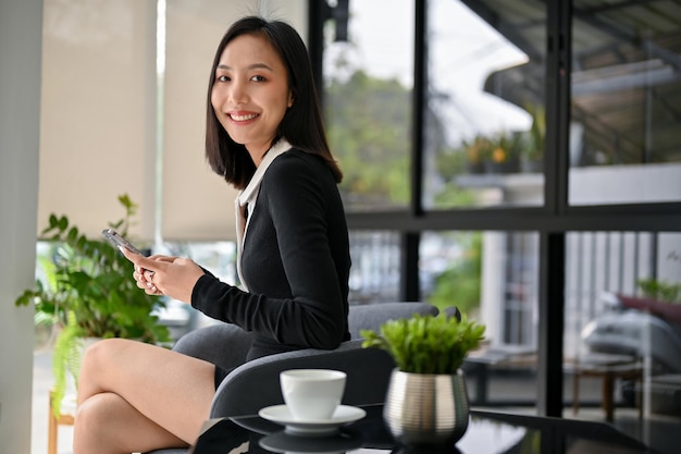 Donna di affari asiatica attraente che tiene il suo telefono che esamina la macchina fotografica e che si rilassa al caffè