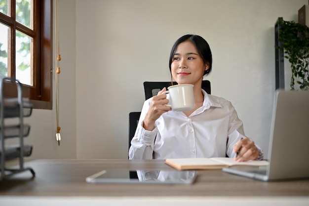 魅力的なアジアのビジネスウーマンが机で仕事をしている間にコーヒーを飲んでいます