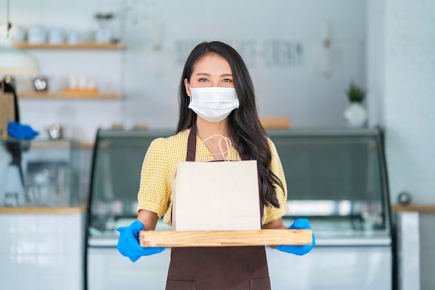 Attractive asian female cafe worker wears face mask and gloves giving takeaway food bag to customerwaitress holding takeout order standing in coffee shop front door entrance new normal lifestyle