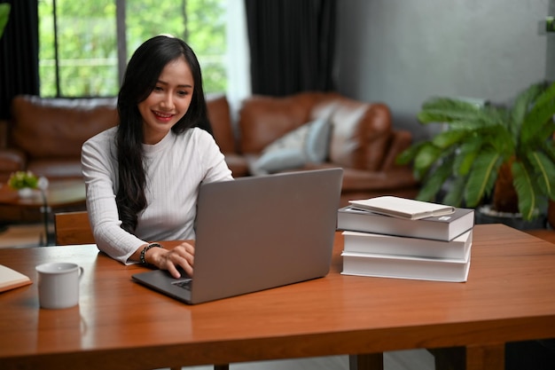 Studentessa di college femminile asiatica attraente che lavora nel suo soggiorno usando il computer portatile