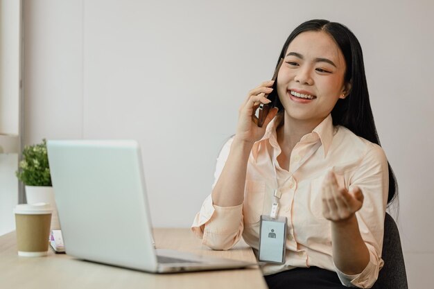 Attractive Asian female employee talking on mobile phone and using laptop at desk