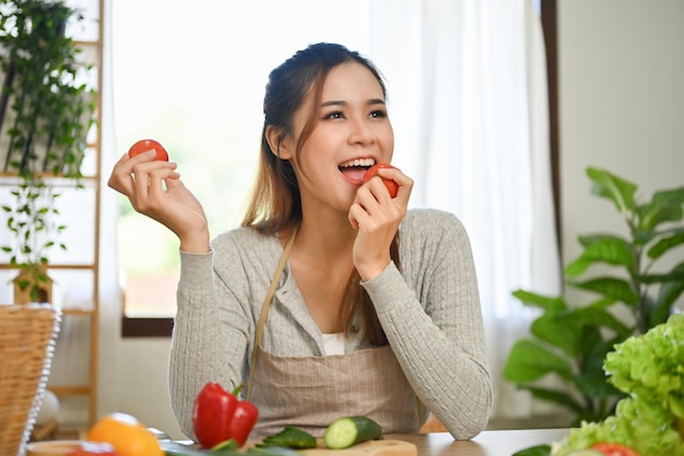 매력적인 아시아 여성은 부엌에서 샐러드 그릇을 만드는 동안 신선한 토마토를 먹는 것을 즐깁니다.
