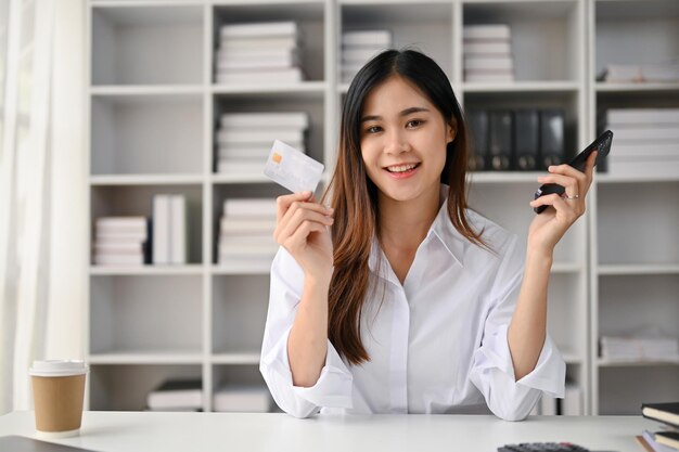Attractive Asian female office employee holding her credit card and smartphone