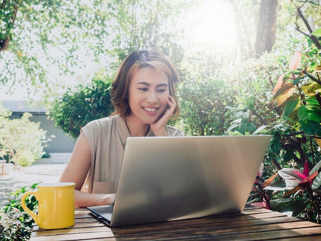 Attractive Asian female portrait with short hair sitting with a hand under chin enjoy with coffee and looking at the laptop computer screen with a relaxing smile in the outdoor garden