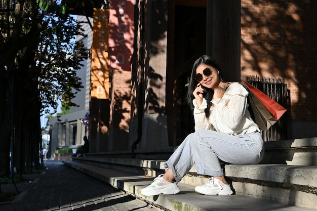 Attractive Asian female traveler with sunglasses and shopping bags sitting on street stairs