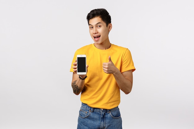 Attractive asian guy in yellow stylish t-shirt with tattoos, showing thumbs-up and phone display as offering customer download amazing app, editing application, game or corporate page