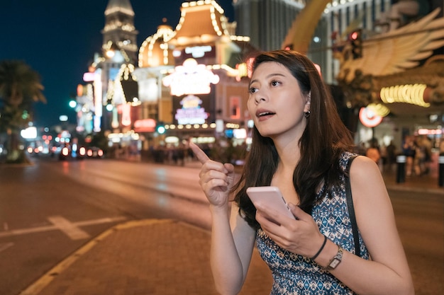 Attractive asian korean female tourist walking through night city street with mobile phone in hands. Smiling girl browsing online maps application using smartphone and searching for direction on road