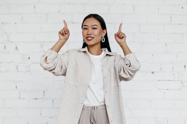 Attractive Asian lady in beige cardigan points fingers up at place for text