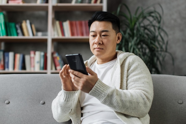 Attractive asian man in casual wear sitting on comfy couch with\
modern smartphone in hands