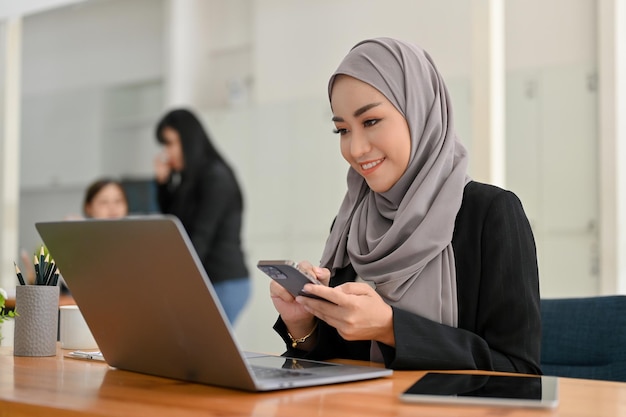 Attraente donna d'affari musulmana asiatica che indossa l'hijab utilizzando lo smartphone e guardando lo schermo del laptop