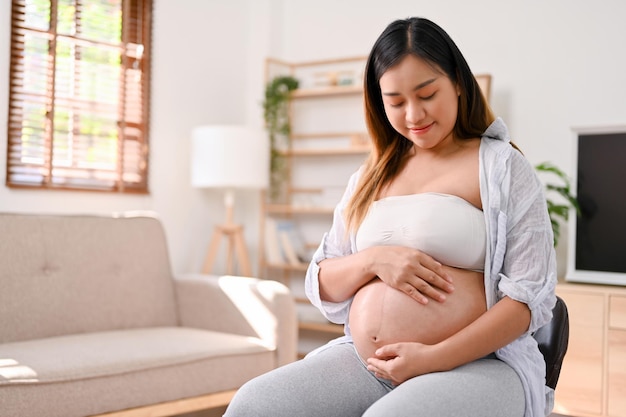 Attractive Asian pregnant woman sits on a chair in the living room touching her belly