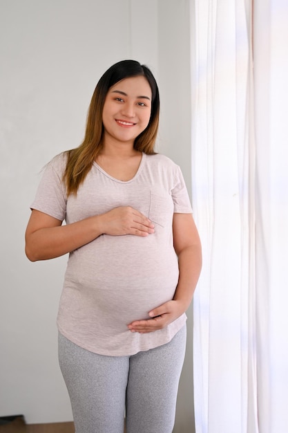 魅力的なアジアの妊娠中の女性が窓のそばに立ち、愛情と気遣いを持ってお腹に触れる