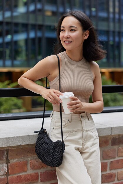 Attractive asian woman holds coffee to go wears t shirt and\
jeans carries small bag looks pensively away enjoys spare time in\
city waits for someone drinks aromatic beverage lifestyle\
concept