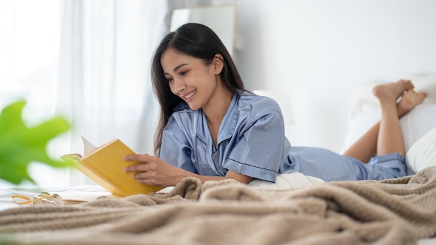 An attractive Asian woman in pajamas enjoys reading a book on her bed in the bedroom in the morning