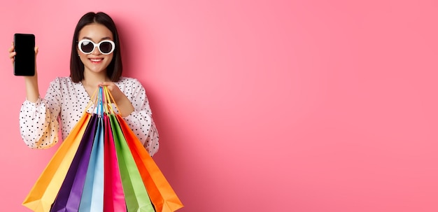 Photo attractive asian woman showing smartphone app and shopping bags buying online via application standi