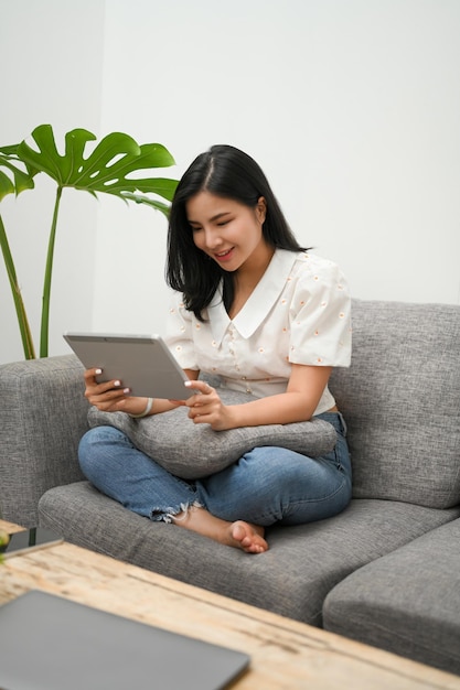 Attractive Asian woman spend her weekend at home relaxes on sofa enjoys using tablet