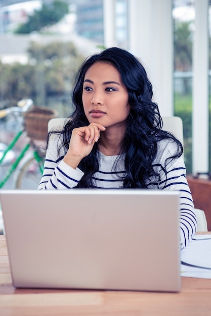 Attractive Asian woman using laptop with hand on chin in office