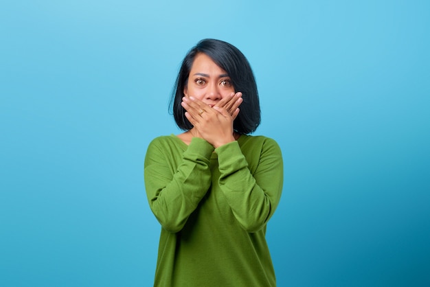 Привлекательная азиатская женщина в повседневной одежде шокирована, прикрывая рот рукой на синем фоне