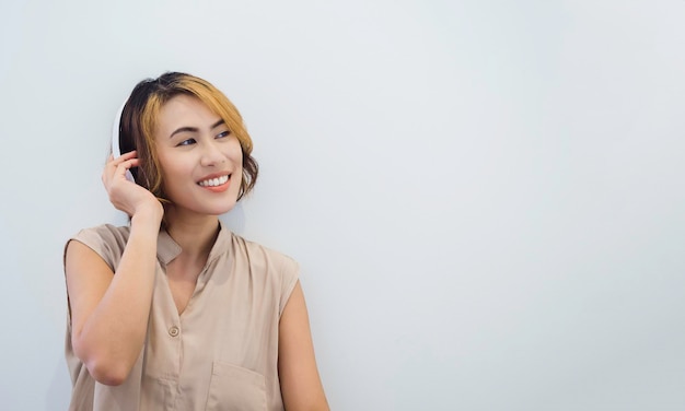 베이지색 민소매 셔츠에 짧은 머리를 한 매력적인 아시아 여성은 복사 공간이 있는 흰색 배경에 해피 스마일이 있는 흰색 헤드폰으로 라디오에서 음악 팟캐스트나 노래를 듣는 것을 즐깁니다.