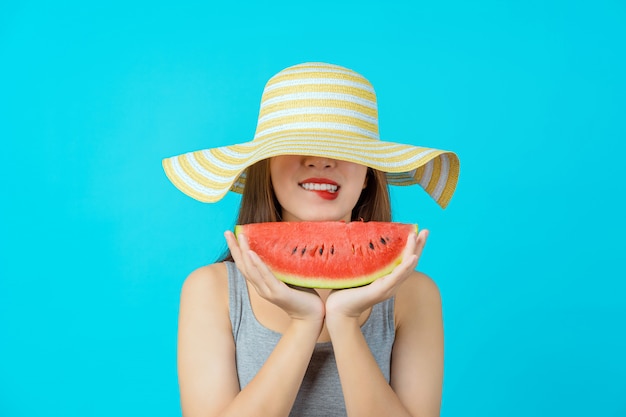 매력적인 아시아 젊은 여자 여름 모자를 착용하고 격리 된 파란색 벽, 섹시한 입에 물린, 복사 공간 및 스튜디오, 패션 여행 및 관광 개념에 수박 조각 슬라이드를 들고