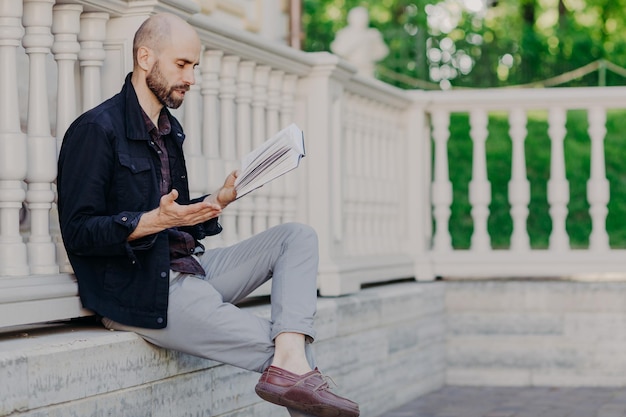 Attractive bald man reads attentively interesting book gestures with hand has stunning plot tries understand feeling of main characters poses outdoor enjoys fresh air and wonderful view