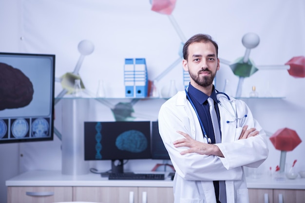 Attractive bearded doctor with arms crossed looking at the camera in his hospital cabinet. Young surgeon in the cabinet.