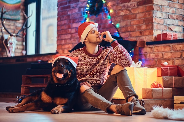 魅力的なあごひげを生やしたヒップスターの男性は、クリスマスの装飾が施された部屋で彼のロットワイラー犬と一緒に床に座っています。